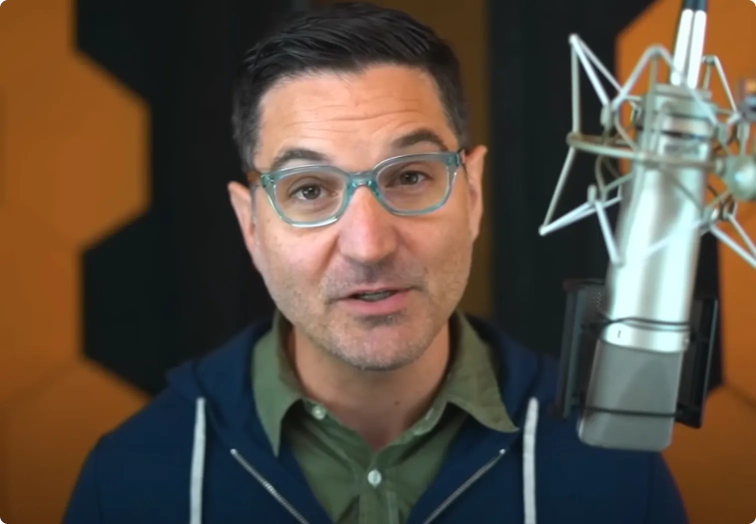 Man with short dark hair and glasses speaking into a professional microphone with orange and black soundproof panels in the background.